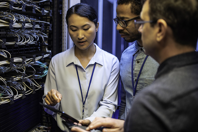 Three IT technician looking at a digital tablet and talking while standing next to a server in a data center.